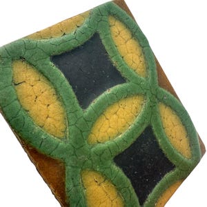 May include: Square ceramic tile with a geometric design. The tile features a pattern of interlocking green and yellow shapes, with black squares at the center. The edges are a brown color.