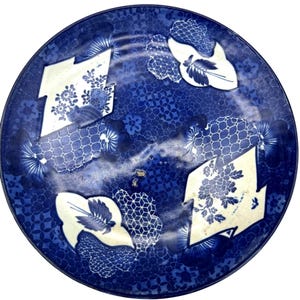 May include: Round blue and white ceramic plate with a floral and geometric pattern. The plate features a dark blue background with a repeating floral design. White, irregular shapes with floral and leaf motifs are scattered across the surface.