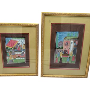 May include: Two framed miniature paintings with gold ornate frames. The paintings depict scenes with figures in colorful clothing, set against a backdrop of buildings and landscapes. The artwork is framed with a cream-colored mat.