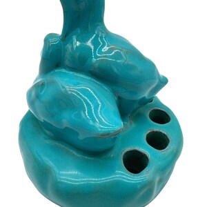 May include: A turquoise ceramic toothbrush holder with a unique, abstract design. The holder features three circular openings and a sculpted form resembling a stylized bird or figure. The glossy finish enhances the vibrant color.