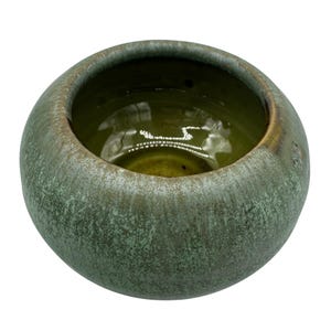 May include: A small, round ceramic bowl with a textured, mottled green exterior and a smooth, olive-green interior. The rim has a subtle brown glaze. The bowl's shape is simple and organic.