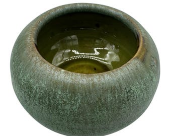 May include: A small, round ceramic bowl with a textured, mottled green exterior and a smooth, olive-green interior. The rim has a subtle brown glaze. The bowl's shape is simple and organic.