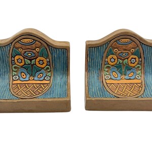 May include: Pair of decorative bookends. Each bookend features a floral design in a basket, with blue, orange, and green accents. The background has a blue, textured pattern. The bookends are made of a tan-colored material.