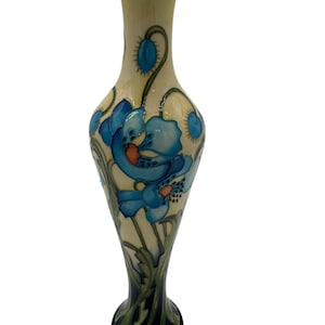 May include: A tall, slender ceramic vase with a cream-colored background and a floral design featuring blue poppies with green stems and leaves. The vase has a flared base and a narrow neck.