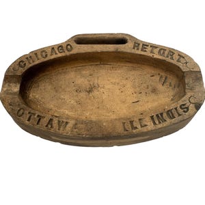 May include: A vintage, oval-shaped ashtray made of a light brown material. The ashtray has a recessed bowl with a small handle and the words "Chicago", "Retort", "Ottawa", and "Ill. Indis" are embossed around the rim.