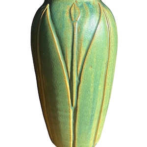 May include: A ceramic vase with a tapered cylindrical shape and a matte green glaze. The surface features vertical, raised relief lines that form stylized, pointed leaf or petal patterns extending from the base toward the narrow opening.