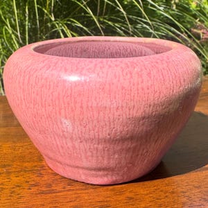 Könnte beinhalten: Eine runde Keramikschale mit weiter Öffnung und einem dicken, abgerundeten Rand. Die Außenseite ist mit einer texturierten, gesprenkelten rosa Glasur mit feinen vertikalen Streifen und einem glänzenden Finish versehen, das eine einheitliche Farbe aufweist.