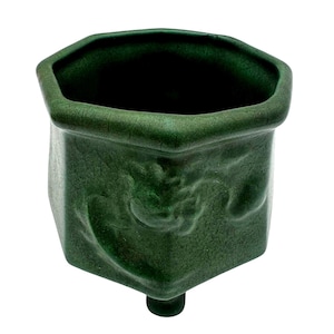 Può includere: Un vaso di ceramica verde scuro con una parte superiore esagonale e una base quadrata. Il vaso ha una superficie strutturata con un sottile disegno in rilievo e un unico piede arrotondato.