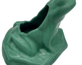 May include: A green frog-shaped planter with a textured surface. The frog is depicted in a sitting position, with a large opening on its back for planting. The planter is designed to resemble a frog sitting on a lily pad.