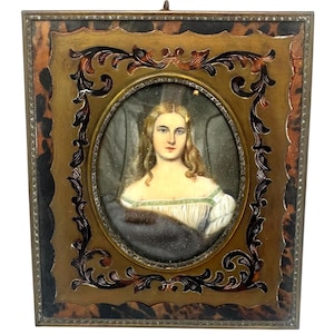 May include: A rectangular portrait miniature featuring a painting of a person with long blonde hair, wearing a white off-the-shoulder garment with green trim and a dark wrap. The portrait is set in an oval frame within a larger, ornate gold and tortoiseshell-patterned rectangular frame.
