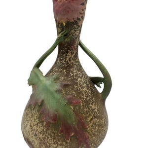 Könnte beinhalten: Eine Keramikvase mit einem gesprenkelten braunen Überzug und grünen und rosa floralen Akzenten. Die Vase hat zwei Griffe, die wie Blätter geformt sind.