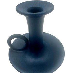 May include: A matte dark blue ceramic candle holder with a wide circular base, a central cylindrical stem with a flared top opening, and a small circular handle attached to the side of the base for easy carrying and handling.