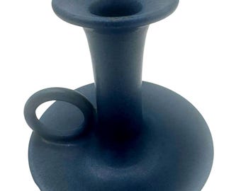May include: A matte dark blue ceramic candle holder with a wide circular base, a central cylindrical stem with a flared top opening, and a small circular handle attached to the side of the base for easy carrying and handling.