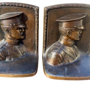 May include: Pair of bronze bookends featuring a relief of a military officer in uniform and hat. The officer has a mustache and the letters "US" are visible on the uniform. The bookends have a decorative border and a curved base.