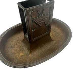 Puede incluir: Un antiguo portafósforos de bronce con base ovalada y un soporte rectangular para la caja de cerillas. El soporte de la caja de cerillas presenta un diseño geométrico. La pieza tiene un aspecto vintage y envejecido.