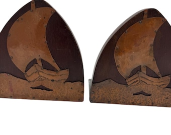 May include: A pair of bookends made of wood and copper. Each bookend features a stylized sailboat with a copper sail and hull. The bookends have a dark brown wood base and are decorated with copper rivets.