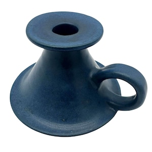 May include: A dark blue ceramic candle holder with a wide, conical base and a circular top platform featuring a central opening. The object includes a small, curved handle attached to the side of the base, finished in a matte, uniform blue glaze.