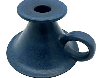 May include: A dark blue ceramic candle holder with a wide, conical base and a circular top platform featuring a central opening. The object includes a small, curved handle attached to the side of the base, finished in a matte, uniform blue glaze.