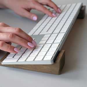 Peut inclure: Un clavier sans fil blanc avec un support en bois. Le clavier est surélevé sur le support, qui est en bois foncé. Le clavier possède un pavé numérique et un pavé tactile.