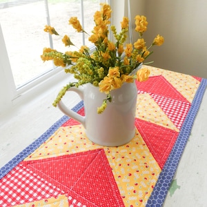May include: A white pitcher filled with yellow flowers sits on a red, yellow, and blue patchwork table runner. The runner has a gingham pattern and a blue border.