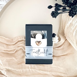 Könnte beinhalten: Eine kleine, dunkelgraue Schachtel mit einem Fenster, das einen weißen Engelsausschnitt und ein herzförmiges Detail zeigt. Die Schachtel ist mit einem hellblauen Band mit dem Wort "Trostlicht" umwickelt. Die Schachtel liegt auf einem beigen Stoff.