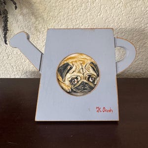May include: A light blue wooden cutout shaped like a watering can, featuring a circular hand-painted portrait of a tan pug with black facial markings. The name K. Oanh is written in red script in the lower right corner of the blue surface.