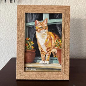 May include: An oil painting in a light brown wood frame depicting an orange tabby cat with white markings standing on a windowsill. The cat is positioned between two terracotta pots containing small orange flowers, with a window and curtains behind.