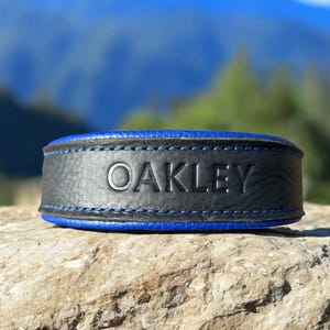 May include: A black leather collar with vibrant blue edges and blue stitching. The name OAKLEY is embossed in capital letters across the center of the collar. The design features a textured surface and a bold, contrasting color scheme.