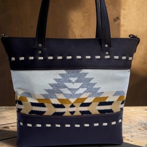 May include: A tote bag with a geometric pattern in shades of gray, yellow, and navy blue. The top and bottom sections are navy blue with white stitching. The bag has dark brown leather handles and trim.