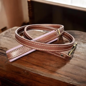 May include: A brown leather dog leash featuring a light lavender padded underside and white contrast stitching. The leash includes a brass-toned metal swivel snap hook at one end and a matching brass-toned metal ring attached to the leather strap.