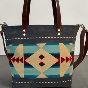May include: A tote bag featuring a central panel with a geometric pattern in turquoise, beige, red, and dark blue. The bag has dark gray leather trim at the top and bottom with white stitching, brown leather handles, and a detachable shoulder strap.