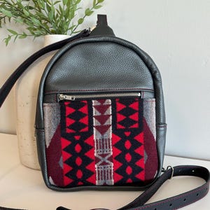 May include: A small black leather backpack with a red, black, and gray patterned fabric panel. The backpack has a single adjustable strap and a zippered pocket on the front.