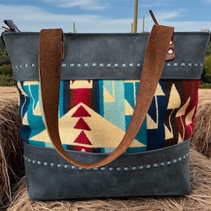 May include: A large tote bag with a Southwestern design. The bag features a gray leather top and bottom with brown leather straps. The center panel has a colorful geometric pattern in red, blue, and cream. The bag is sitting on a bed of hay.