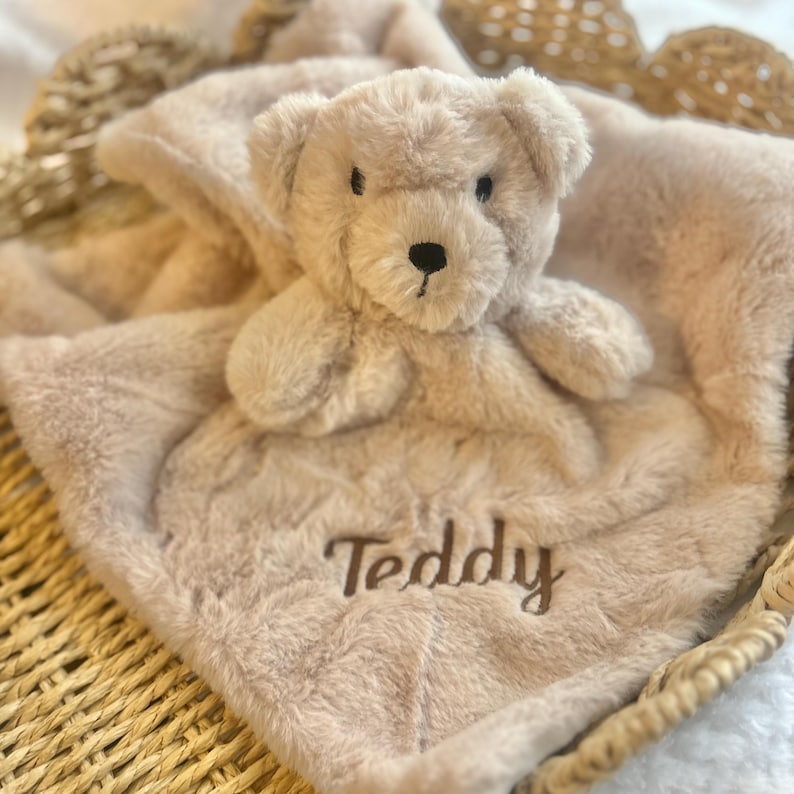 May include: A soft, beige teddy bear plush toy attached to a matching blanket. The word "Teddy" is embroidered on the blanket. The blanket and bear are nestled in a woven basket.