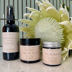 May include: A selection of skincare products from The Herbal Estate. The image shows a black spray bottle of Mugwort and Rose Toner, a jar of Organic Tallow Balm, and a jar of Facial Polish. A large, pale green flower is in the background.