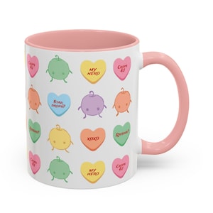 May include: White ceramic mug with a pink interior and handle. The mug is decorated with colorful heart-shaped candies, each featuring a cute character and a short message like "XOXO" and "My Hero".