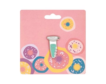 May include: A white and green enamel pin shaped like a test tube with a blue liquid inside. The pin is on a pink card with a white border and colorful circles.