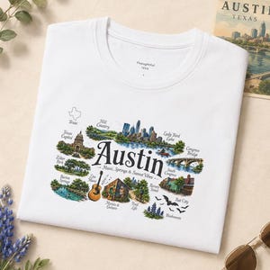 May include: A white t-shirt featuring a colorful graphic illustration of Austin, Texas landmarks including the Capitol, Congress Bridge, and Lady Bird Lake, with the text "Austin Music, Springs & Sunset Vibes" and various labels like "Zilker Park" and "Bat City".