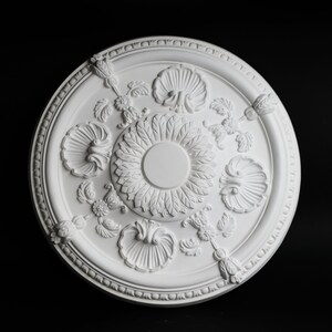 May include: White ceiling medallion with a decorative floral design. The medallion has a raised center with a circular opening and is surrounded by a border of small beads.
