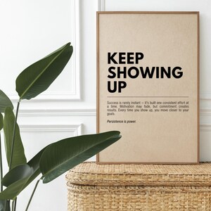 Könnte beinhalten: Ein gerahmter Druck mit den Worten "KEEP SHOWING UP" in fetten schwarzen Buchstaben. Darunter erläutert kleinerer Text die Bedeutung von konsequenter Anstrengung und Engagement. Der Druck befindet sich auf einem hellbraunen Hintergrund und liegt auf einem geflochtenen Korb.