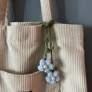 May include: A cream-colored corduroy tote bag with a front pocket and a green crocheted floral charm. The charm features two light blue crocheted flowers with pearl centers, adding a delicate touch to the bag.