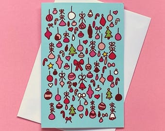 May include: A greeting card with a light blue background featuring a pattern of Christmas ornaments in red, pink, and green. The card is on top of a white envelope, set against a pink background.