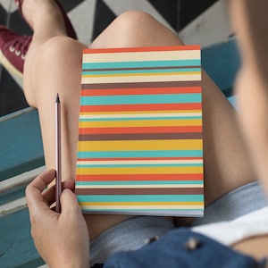 Peut inclure: Une personne assise sur un banc bleu tenant un carnet à couverture rayée colorée et un crayon.