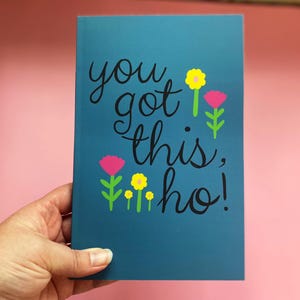Puede incluir: Un cuaderno rectangular de color verde azulado con el texto cursivo negro "you got this, ho!" rodeado de pequeñas ilustraciones coloridas de flores amarillas y rosas con tallos verdes. La cubierta tiene un acabado mate y un diseño plano.