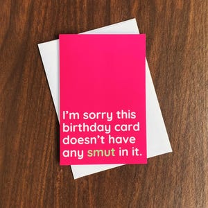 Op de afbeelding: Een felroze verjaardagskaart met de tekst "I'm sorry this birthday card doesn't have any smut in it." De kaart ligt op een witte envelop, tegen een donkere houtnerf achtergrond.