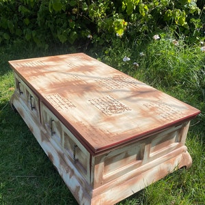 May include: A distressed white coffee table with a red trim and two drawers. The top of the table is painted with a brown stain and has a decorative stencil design.