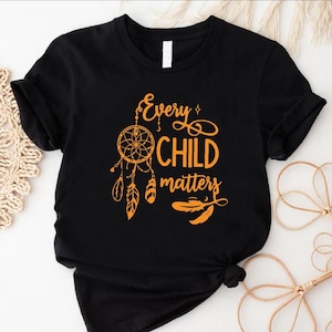 May include: Black t-shirt with the text "Every Child Matters" in orange script, featuring a dreamcatcher and feathers design. The shirt is a classic crew neck style.