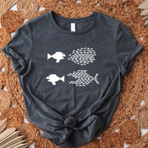 May include: A dark gray t-shirt with a white graphic of two fish. The fish on the left is facing right and has a school of fish coming out of its mouth. The fish on the right is facing left and has a school of fish following behind it.