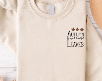 May include: A cream-colored sweatshirt with the text "Autumn Breeze & Beautiful Leaves" in black. Above the text are three brown maple leaf graphics. The sweatshirt has a crew neck and long sleeves.