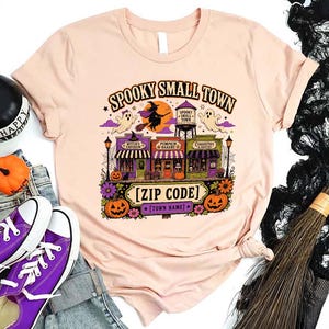 May include: A light peach t-shirt featuring a colorful graphic of a Halloween-themed town with the text "SPOOKY SMALL TOWN", "[ZIP CODE]", and "[TOWN NAME]", depicting a witch on a broom, ghosts, a water tower, and storefronts.
